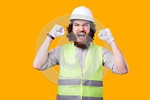 An upset young engineer is looking at the camera screaming with both hands up near a yellow wall