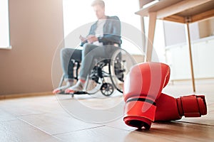 Upset young ex-sportsman with disability and inclusiveness sitting on wheelchair. Boxer`s gloves lying on floor. Can`t photo