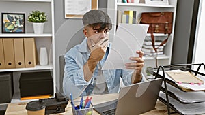 Upset young hispanic business worker, drowned in paperwork, reading a distressing document on his laptop at the office