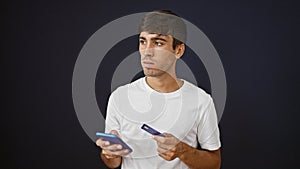 Upset young hispanic man looking serious whilst shopping online, holding credit card and typing on smartphone. isolated against