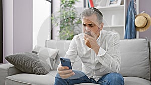 Upset young hispanic man, sporting grey hair and a beard, engrossed in a serious smartphone ordeal while comfortably nestled on