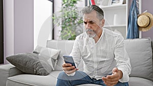 Upset young hispanic man, sporting grey hair and a beard, engrossed in a serious smartphone ordeal while comfortably nestled on