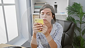 Upset young hispanic woman business worker holds smartphone in office, doubting her professional success, showcasing sadness