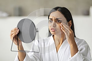 Upset young indian woman looking at mirror, touching her cheeks