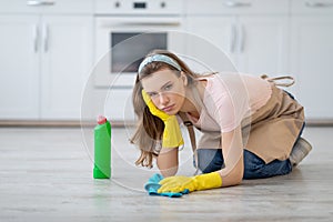 Upset young lady in rubber gloves and apron washing floor at kitchen, disliking house chores, feeling tired and unhappy