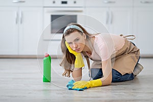 Upset young lady in rubber gloves and apron washing floor at kitchen, disliking house chores, feeling tired and unhappy photo