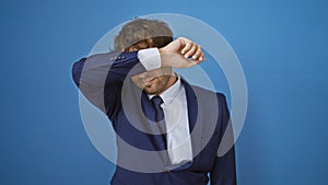 Upset young man in business suit hides face with arm, embodying sightless denial concept on isolated blue backdrop