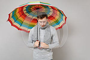 Upset young man in gray sweater, scarf looking down, holding colorful umbrella on grey background. Healthy