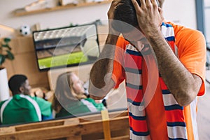 upset young man in orange t-shirt and scarf holding hands on head while his friends watching soccer match on tv screen