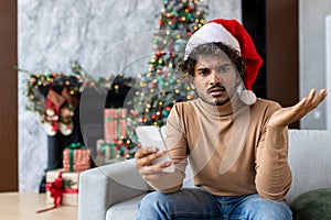 Upset young man sitting on sofa at home in red santa hat, hispanic looking disappointed at camera holding phone, got bad