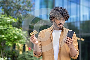 Upset young muslim man standing on city street, holding credit card and looking at phone screen in frustrated, worried