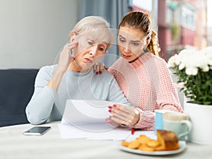 Upset young woman with aged mother looking worriedly at papers at home