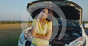 Upset young woman driver talking angrily on cell phone with assistance service near a broken car with open car hood on a