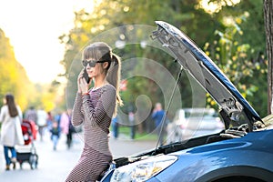 Upset young woman driver talking angrily on cell phone with assistance service near a broken car with open hood on a city street