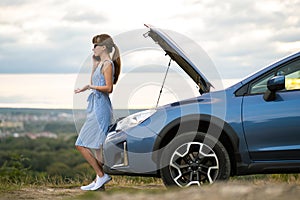 Upset young woman driver talking angrily on cell phone with assistance service near a broken car with open hood while inspecting