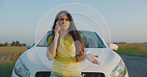 Upset young woman driver talking angrily on cell phone with assistance service near a broken car on a road. Waiting for