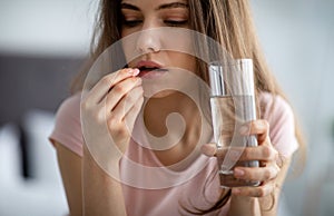 Upset young woman with glass of water taking pill, not feeling well, using medical drug at home