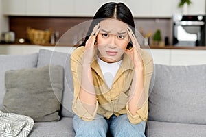 Upset young woman has strong headache, sitting on sofa at home and massaging temples, feels tired and exhausted