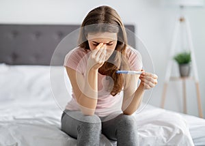 Upset young woman holding pregnancy test, touching her face in stress on bed at home