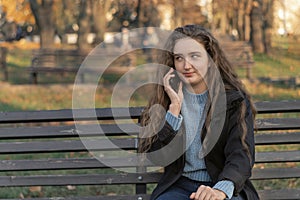 Upset young woman with long wavy brown hair sits on bench in park talks on phone. Woman in autumn park