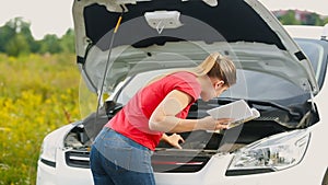 Upset young woman looking at the car manual and trying to repair brakedown vehicle on the deserted road