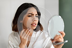 Upset young woman looking at mirror and touching her face