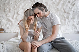 Upset young woman with negative pregnancy test crying on bed, loving husband supporting her at home
