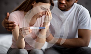 Upset young woman showing negative pregnancy test, her black husband supporting her in bedroom, selective focus