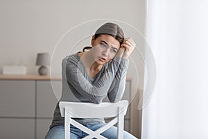 Upset young woman sits alone, looking away with sad expression at home interior