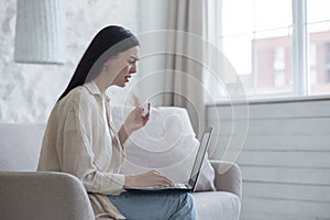 Upset young woman sits on the couch with a laptop. Crying, wiping tears with a napkin