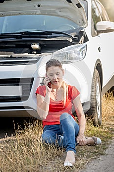 Upset young woman sitting on ground next to broken car calling car service for help