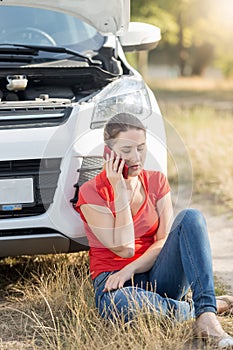 Upset young woman sitting at her broken car in field and calling tow truck for help