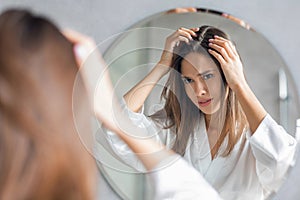 Upset Young Woman Standing Near Mirror And Looking At Her Hair Roots