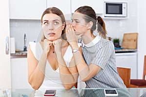 Upset young women whispers in the ear and talking at table in home