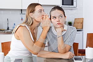 Upset young women whispers in the ear and talking at table in home