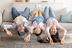 Upside down portrait of happy young family lying on sofa with heads down. Mother, father and daughter having fun