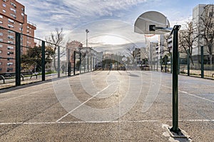 An urban basketball court with rough cement floors, a perimeter with metal fences and baskets on metal posts screwed to the ground