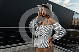 Urban elegant young woman fashion model in fashionable straw white hat in vintage shirt rests in city near modern black building.