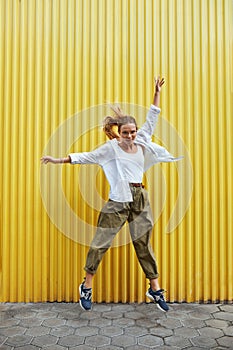 Urban. Fashion Girl Motion Portrait. Sexy Model In Casual Clothes And Glasses Against Yellow Iron Fence. Beautiful Woman Jumping