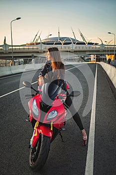 Urban fashion. Outdoor portrait of pretty young woman in black leather jacket sitting on red motobike on sunset