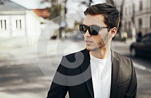 Urban fashion portrait of young handsome businessman in casual suit in dark sunglasses sitting in the street goind to the work and