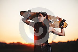 Urban fashionable girl with longboard posing outdoors on the road at sunset.
