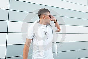 Urban handsome young hipster man in a trendy white t-shirt in stylish sunglasses with fashionable hairstyle posing near a modern