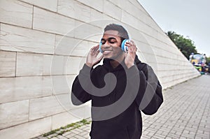 urban happy smiling african man in wireless headphones enjoying listening to music wearing black hoodie on city street