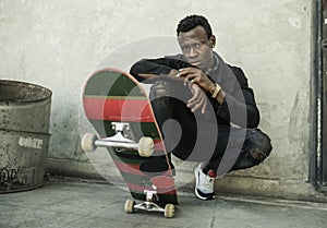 Urban portrait of young attractive and serious black afro American man with skate board squatting on street corner looking cool