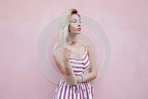 Urban pretty young blonde woman in a summer stylish striped sundress rests near a pink vintage wall in the city. Cute urban girl