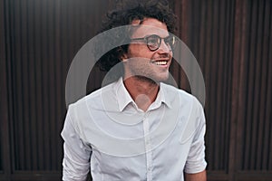 Urban shot of handsome young businessman with glasses, smiling, posing outdoors. Male student posing against brown wall. Smart guy