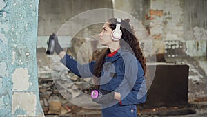 Urban street artist in headphones is listening to music and painting on the wall in old empty house. Pretty girl is