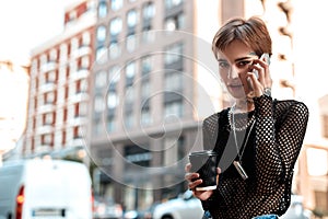 Urban Style. Young stylish woman standing outdoors with cup of coffee talking on phone smiling curious