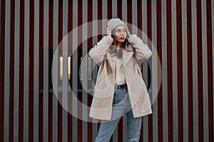 Urban stylish young woman in a fashionable eco coat in vintage blue stylish jeans straightens trendy gray knitted hat near a